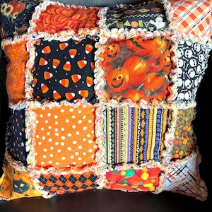 May include: A square rag quilt pillow featuring a patchwork design of various Halloween-themed fabrics. The squares display patterns of pumpkins, candy corn, skulls, polka dots, and stripes in orange, black, white, and purple colors.