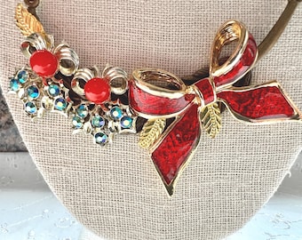 Puede incluir: Un collar vintage con un gran lazo rojo, ribetes y detalles dorados. El collar también incluye racimos de pedrería azul-verdosa iridiscente y orbes rojos. El diseño se presenta sobre un fondo neutro.