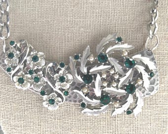 May include: Silver-toned statement necklace with floral and leaf designs. The necklace features clusters of small, green gemstones and faux pearls. The chain is a chunky, silver link style, creating a bold, vintage aesthetic.