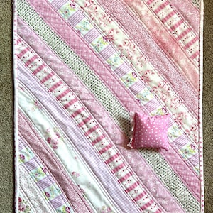 May include: A rectangular quilt featuring diagonal strips of fabric in various shades of pink, white, and green. The patterns include floral prints, stripes, polka dots, and small geometric shapes. A small, square pink polka-dot pillow sits on top.