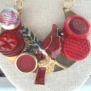 May include: A unique necklace featuring a red cardinal bird design with gold accents, surrounded by various red buttons and a clear iridescent button. The necklace is set on a gold-toned metal base.