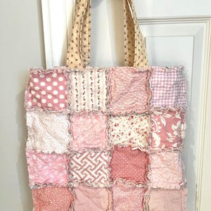 May include: A square tote bag constructed from sixteen patchwork fabric squares in various shades of pink and white. The squares feature diverse patterns including polka dots, stripes, and floral prints, joined with frayed, exposed seams and two matching handles.