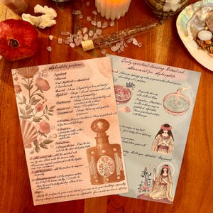 May include: Two illustrated pages with text and drawings on a wooden surface. One page details an Aphrodite perfume recipe, the other a spiritual cleansing ritual. The pages are adorned with floral and botanical illustrations.