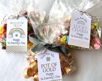 May include: Three clear plastic bags filled with different candies and snacks. Each bag has a colorful tag attached with a St. Patrick's Day theme. The tags read "Rainbow Seeds", "Lucky Charm", and "Pot of Gold".