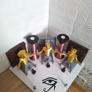 May include: A tabletop display featuring three golden figures with outstretched arms, each on a gray platform. Two tall, copper-colored cylinders with black tops flank the figures. The display includes brown rectangular panels and an eye symbol.