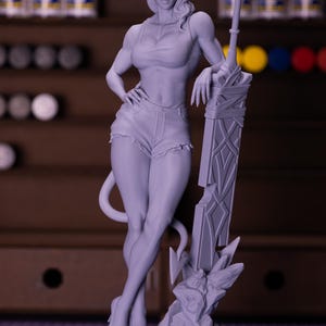 Puede incluir: Figura gris de una demonio femenina con cuernos, cola y espada. Viste camiseta sin mangas, pantalones cortos y tacones. La figura está de pie sobre una base rocosa. El fondo está borroso, con botes de pintura visibles.