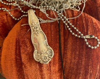 May include: An orange velvet pumpkin with a silver spoon-shaped ornament and a silver beaded chain. The spoon has floral detailing. Dried, light-colored plant material is also present.