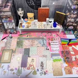 May include: A craft workspace featuring a wide array of decorative paper sheets, floral and botanical stickers, patterned cardstock, and various stationery supplies spread across a pink mat, with organised storage drawers in the background.