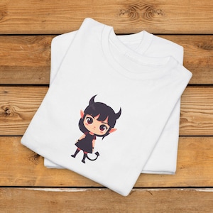 Op de afbeelding: Wit T-shirt met een cartoon duivelafbeelding. De illustratie toont een personage met zwarte hoorns, een zwarte jurk en een staart. Het ontwerp staat op een opgevouwen T-shirt, gestapeld op een ander wit T-shirt. De shirts liggen op een houten oppervlak.