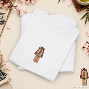 May include: A white t-shirt featuring a small, centered embroidered graphic of a rectangular wooden Japanese omamori amulet. The amulet is light brown with dark red tassels, a red circular emblem, and black Japanese text reading 厄除.