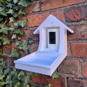 Op de afbeelding: Witte vogelhuisvormige houder voor een beveiligingscamera met een vogel silhouet. Het heeft een dienblad en ruimte voor een Ring-camera. Gemonteerd op een rode bakstenen muur met groene klimop.