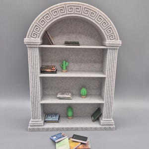 May include: A miniature gray bookshelf with three shelves, decorated with a Greek key design. The shelves hold small books and cactus figurines. A few additional miniature books are scattered in front of the bookshelf.