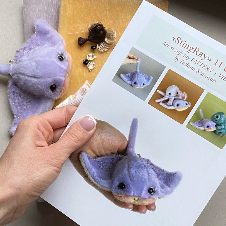 Op de afbeelding: Een lavendelkleurig pluche pijlstaartrog-speeltje met donkere ogen en een lichte onderkant, vergezeld van een gedrukte handleiding getiteld StingRay 11 cm Artist soft toy PATTERN + VIDEO door Tetiana Skalozub, met foto's van het speelgoed.