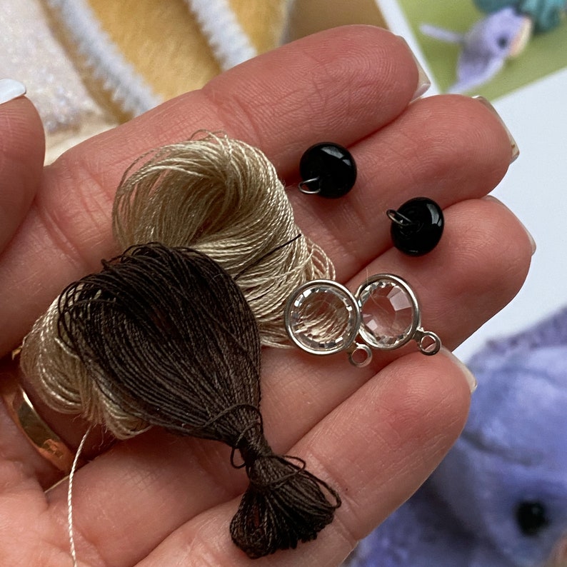 Op de afbeelding: Twee kleine zwarte bolvormige knopen, twee heldere gefacetteerde ronde bedels met zilverkleurige metalen zettingen, en twee kleine bundels borduurgaren in donkerbruin en lichtbeige, die samen in een handpalm rusten.