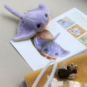 Op de afbeelding: Twee lavendelkleurige pijlstaartrog knuffels met donkere ogen, roze onderkanten en kraalaccenten op de vinnen. De speeltjes liggen naast een geprint instructieblad getiteld Stingray, een stuk gele stof, witte pijpenragers en naaigaren.