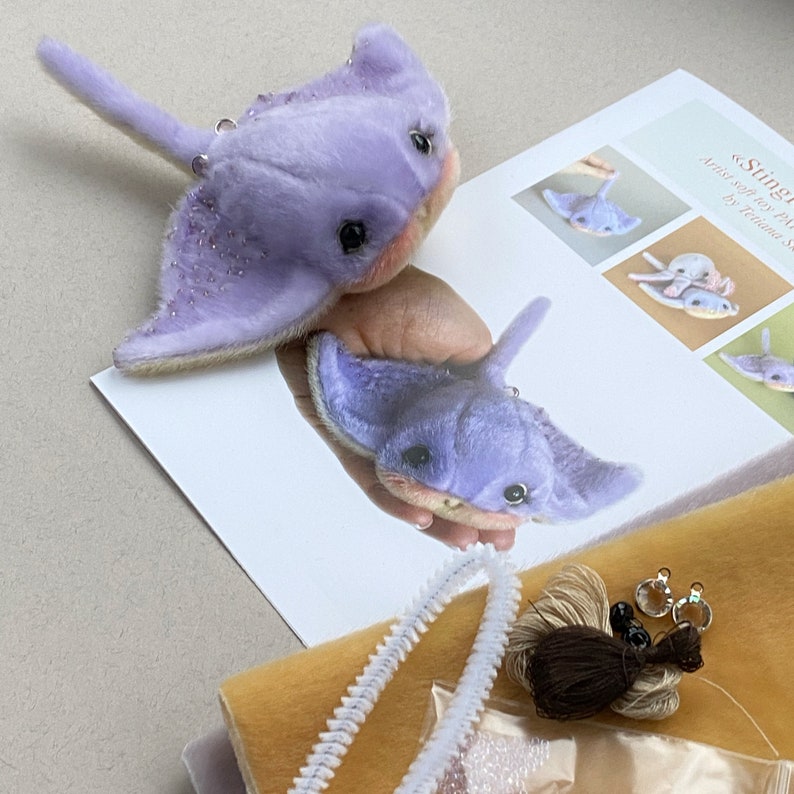 Op de afbeelding: Twee lavendelkleurige pijlstaartrog knuffels met donkere ogen, roze onderkanten en kraalaccenten op de vinnen. De speeltjes liggen naast een geprint instructieblad getiteld Stingray, een stuk gele stof, witte pijpenragers en naaigaren.