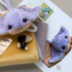 Op de afbeelding: Twee pluche pijlstaartrog-speeltjes gemaakt van zachte lavendelkleurige stof met donkere ogen en roze wangen. E&eacute;n speeltje wordt in een hand gehouden, terwijl de andere op een geel en grijs stoffen staal ligt. Een klein kwastje met zwarte en doorzichtige kralen ligt ernaast.