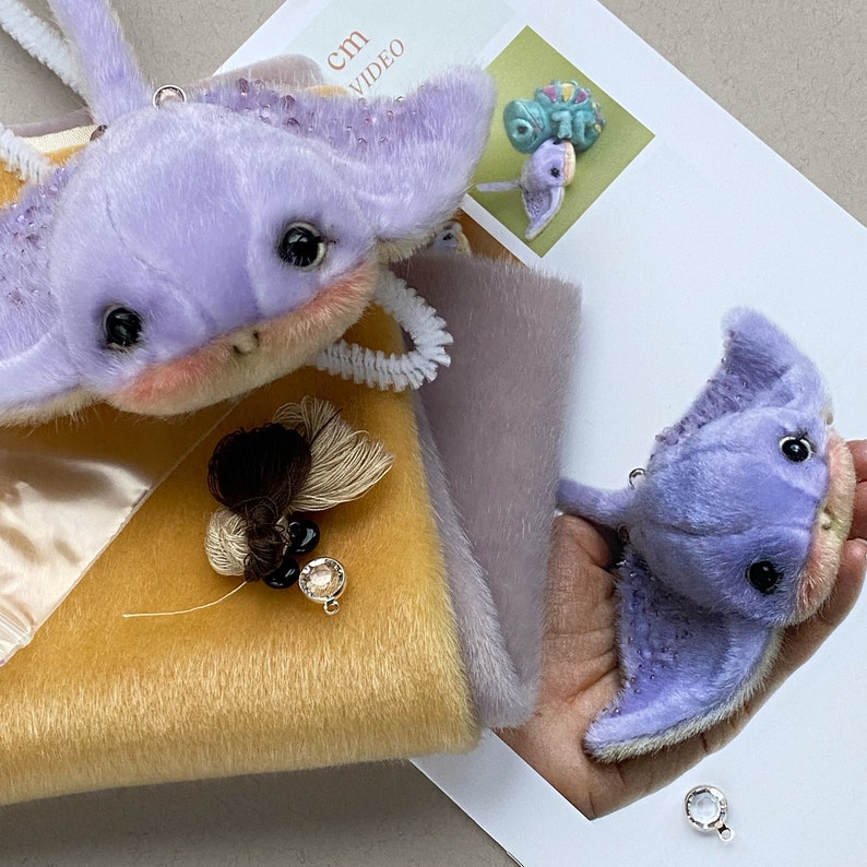Op de afbeelding: Twee pluche pijlstaartrog-speeltjes gemaakt van zachte lavendelkleurige stof met donkere ogen en roze wangen. E&eacute;n speeltje wordt in een hand gehouden, terwijl de andere op een geel en grijs stoffen staal ligt. Een klein kwastje met zwarte en doorzichtige kralen ligt ernaast.