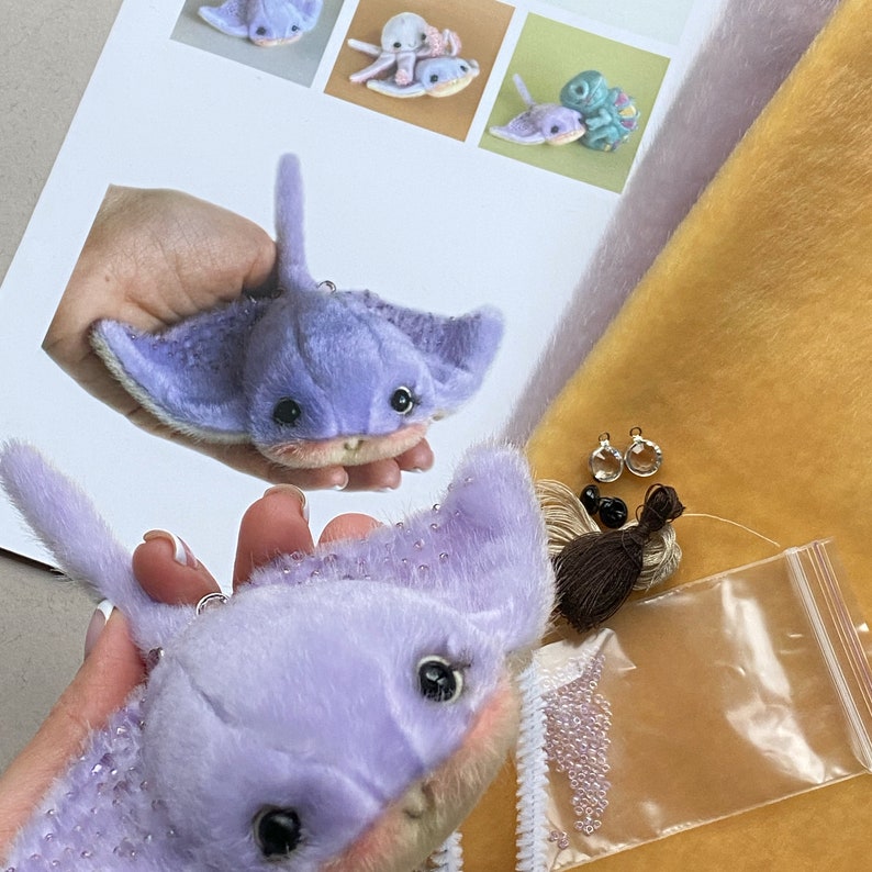Op de afbeelding: Een lavendelkleurige pluche pijlstaartrog met een zachte textuur, donkere ogen en een puntige staart. De set bevat een klein plastic zakje met kleine doorzichtige kraaltjes, twee metalen ringetjes, twee zwarte kraaltjes en een bundel bruin garen.