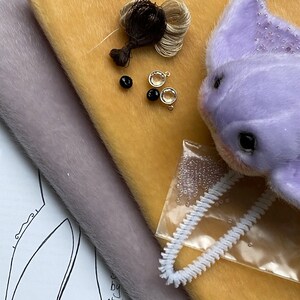 Op de afbeelding: Een lavendelkleurige pluche wezenkop met kraaldetails, geplaatst op mosterdgele en grijze stofstalen. Inbegrepen zijn twee donkerbruine draadbundels, twee zwarte kralen, twee heldere kristallen bedels en een witte pijpenrager in een plastic zakje.