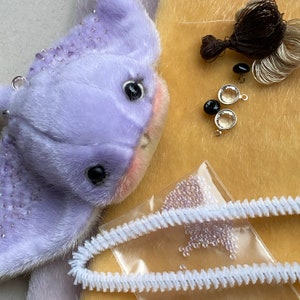 Op de afbeelding: Een lavendelkleurig pluche wezen met donkere ogen en subtiele roze wangen, vergezeld van knutselbenodigdheden waaronder donkerbruin garen, twee zwarte knopen, twee doorzichtige ronde bedels met zilveren randen, een zakje met kleine lavendelkleurige kralen en witte pijpenragers.