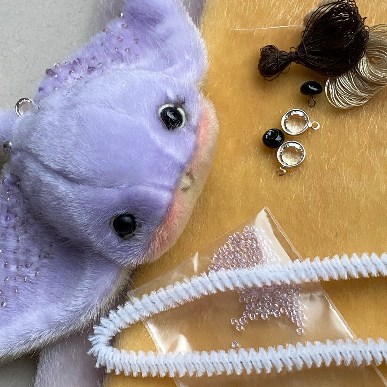 Op de afbeelding: Een lavendelkleurig pluche wezen met donkere ogen en subtiele roze wangen, vergezeld van knutselbenodigdheden waaronder donkerbruin garen, twee zwarte knopen, twee doorzichtige ronde bedels met zilveren randen, een zakje met kleine lavendelkleurige kralen en witte pijpenragers.