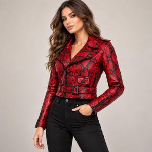 Pode incluir: Um casaco de motociclista com padrão de pele de cobra em vermelho e preto, com gola entalhada, mangas compridas com punhos de fecho, fecho frontal, um pequeno bolso com aba no peito e um cinto a condizer com fivela preta na cintura.