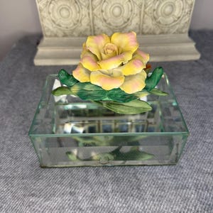 May include: A yellow and pink porcelain rose with green leaves sits on a clear glass mirrored box.