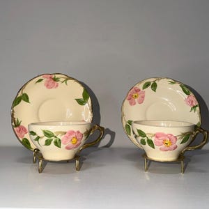 May include: Two matching teacups and saucers with a floral design. The cups and saucers are cream-coloured with pink flowers and green leaves. The rims are trimmed in gold. Each cup sits on a small gold-coloured metal stand.