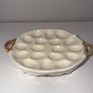 May include: Off-white ceramic escargot dish with 12 oval indentations. The dish has two woven handles and a slightly ruffled edge. A classic serving piece for French cuisine.