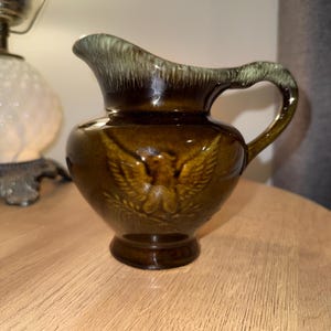 Puede incluir: Una jarra de cerámica marrón con un esmalte verde y un diseño de águila en relieve. La jarra tiene una boca ancha y un asa.