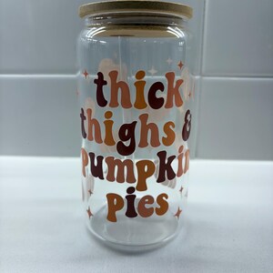 May include: A clear glass can-shaped tumbler with a bamboo lid and a clear straw. The tumbler features the text "thick thighs & pumpkin pies" in shades of orange and brown. Small star graphics are also present.