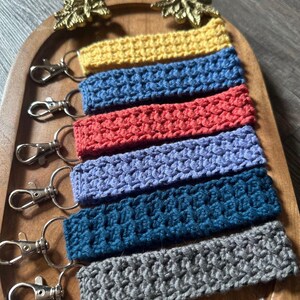 May include: Six crocheted keychains in different colours, including yellow, blue, red, purple, teal, and grey, displayed on a wooden tray with gold accents.