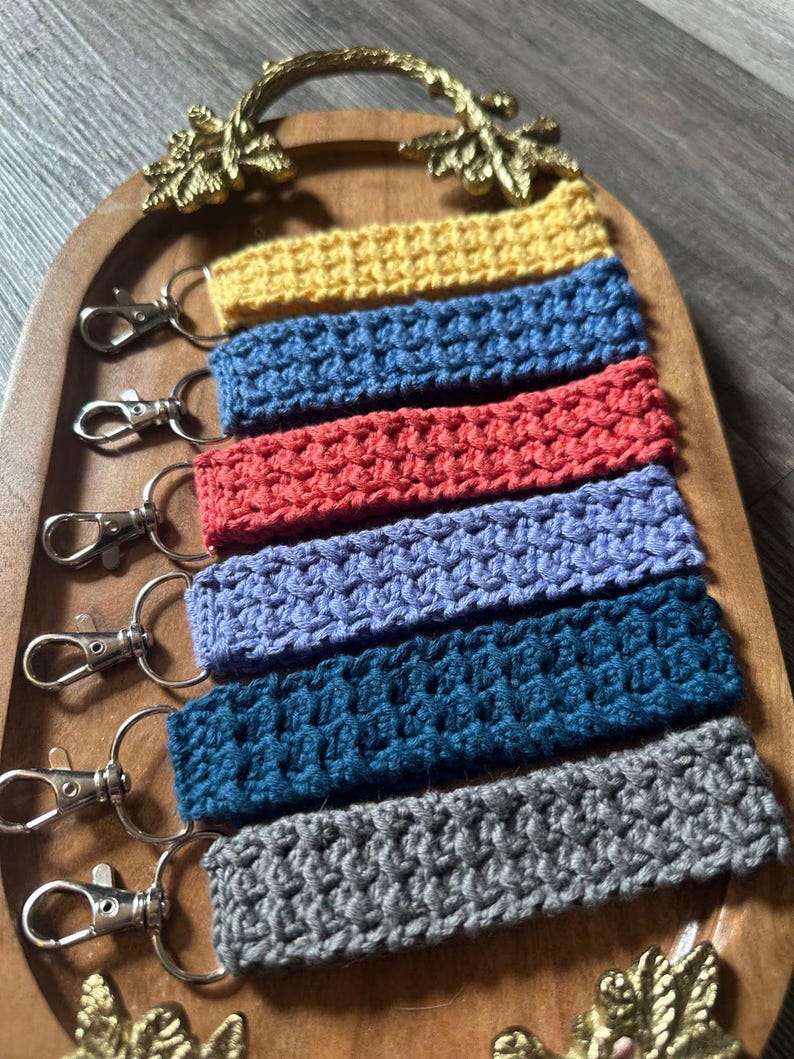 May include: Six crocheted keychains in different colours, including yellow, blue, red, purple, teal, and grey, displayed on a wooden tray with gold accents.