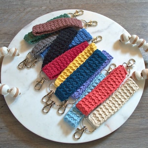 May include: A set of ten crocheted keychains in various colours, including brown, green, grey, black, red, yellow, blue, light blue, coral, and beige. Each keychain has a silver metal key ring.