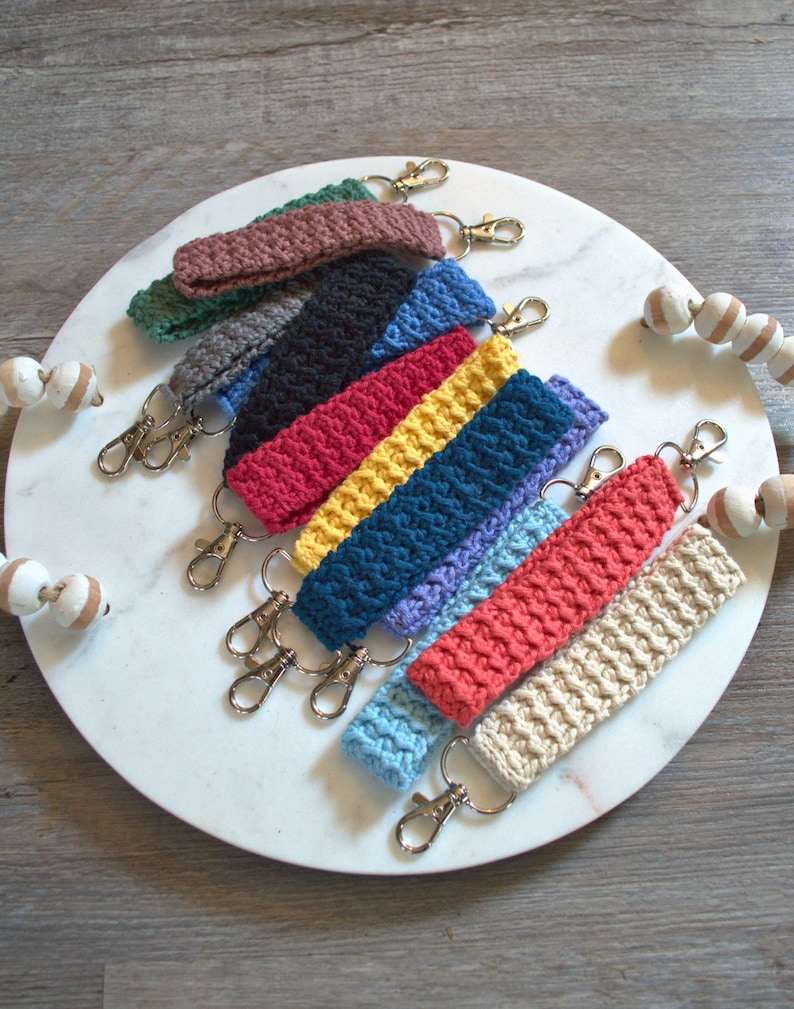 May include: A set of ten crocheted keychains in various colours, including brown, green, grey, black, red, yellow, blue, light blue, coral, and beige. Each keychain has a silver metal key ring.