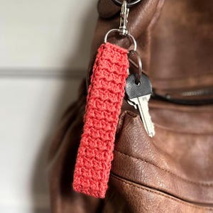 May include: A red crocheted keychain attached to a brown leather bag with silver key rings and keys.