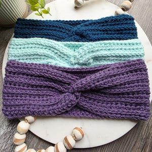 May include: Three handmade crochet headbands in teal, mint green, and purple. The headbands are knotted at the center and have a textured appearance. They are displayed on a white marble-like surface, with decorative wooden beads.