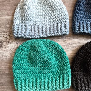 May include: A collection of handmade crochet beanies in various colors. The beanies include shades of light blue, teal, navy, and black. The hats are made with a ribbed brim and a textured stitch pattern. The beanies are displayed on a wooden surface.