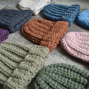 May include: A collection of ten crocheted hats in various colors, including purple, green, brown, pink, blue, and gray. The hats are all made with a chunky yarn and have a simple, ribbed design.