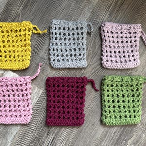 May include: Six crocheted soap sacks in different colors: yellow, gray, pink, burgundy, green, and pink. The sacks are made with a simple stitch pattern and have a drawstring closure.