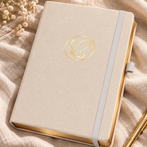 Può includere: Un taccuino con copertina in tessuto beige caratterizzato da un emblema a farfalla in lamina d'oro all'interno di un cerchio sulla copertina. Il taccuino include una fascia elastica di chiusura verticale grigio chiaro, un passante per penna e bordi dorati.