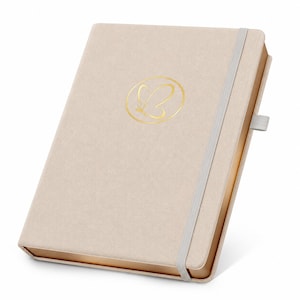 May include: A beige hardback notebook with a textured fabric cover, featuring a gold foil butterfly logo centred on the front. The notebook includes a light grey elastic closure band, a matching pen loop on the side, and metallic gold page edges.