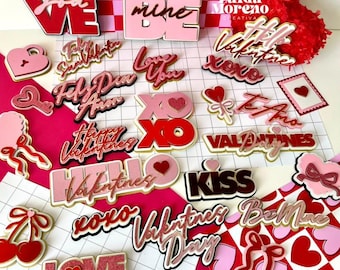Könnte beinhalten: Eine Sammlung von Dekorationen zum Valentinstag in Rot-, Rosa- und Weißtönen. Die Dekorationen umfassen Herzformen, das Wort "LOVE" und andere romantische Phrasen. Die Artikel sind auf einer rosa-weiß karierten Oberfläche angeordnet.