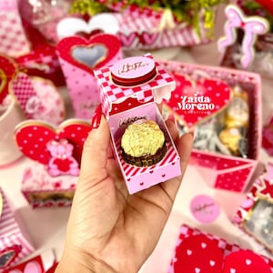 May include: A pink and red Valentine's Day gift box with a gold-wrapped chocolate candy inside. The box has a checkered top and heart details. The image includes other Valentine's Day themed decorations, including hearts and roses.