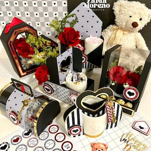 May include: A collection of Valentine's Day gifts, including red roses in decorative boxes, a teddy bear, and heart-shaped stickers. The gifts are arranged on a white surface with a black and white heart pattern.