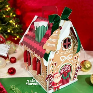 以下が含まれることがあります： クリスマスをテーマにしたジンジャーブレッドハウス型のギフトボックス。赤と白のアクセント、緑のリボン、サンタクロースのフィギュアが特徴です。ボックスには赤い屋根、煙突、ハンドルが付いています。「Zaida Moreno Creativa」の文字が見えます。
