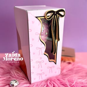 Puede incluir: Una caja de regalo rosa con una ventana, decorada con lazos y corazones. La caja tiene un borde dorado y un lazo negro. El texto "Zaida Moreno Creativa" es visible en el lateral de la caja.
