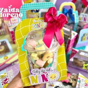 Könnte beinhalten: Eine gelbe Geschenkbox mit einem durchsichtigen Plastikbecher, gefüllt mit bunten Marshmallows. Die Box hat einen Griff und eine große rosa Schleife. Auf der Box steht "Feliz día del NIN" und "Siempre SONRIE".