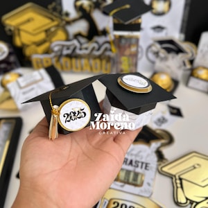 May include: Black graduation cap-shaped gift boxes with gold accents and the year 2025 on a circular tag. The boxes are held in a hand, with other graduation-themed decorations in the background. The text "Zaida Moreno Creativa" is also visible.